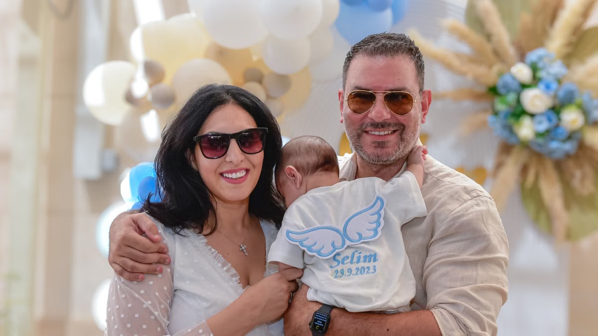 Family holding a baby wearing a personalized outfit, celebrating a special occasion indoors.